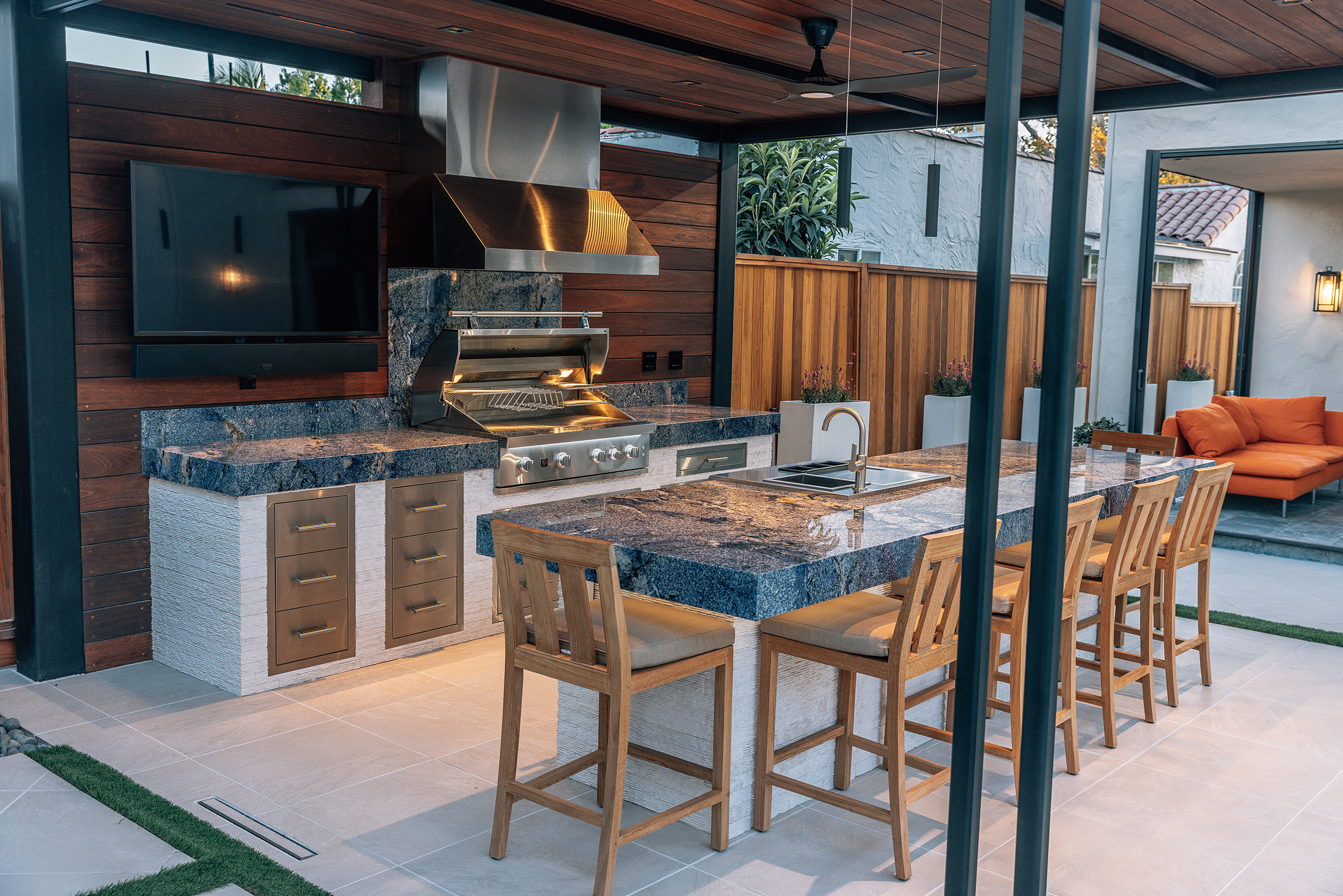 Outdoor kitchen with stainless steel vent hood duct cover over a Summerset grill, stone-top island, and wood cabinet drawers under a covered patio.
